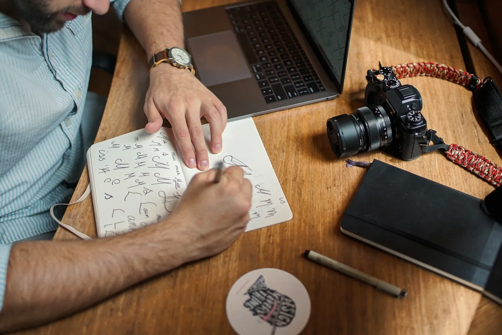 Person writing notes on paper next to a camera on a wooden desk